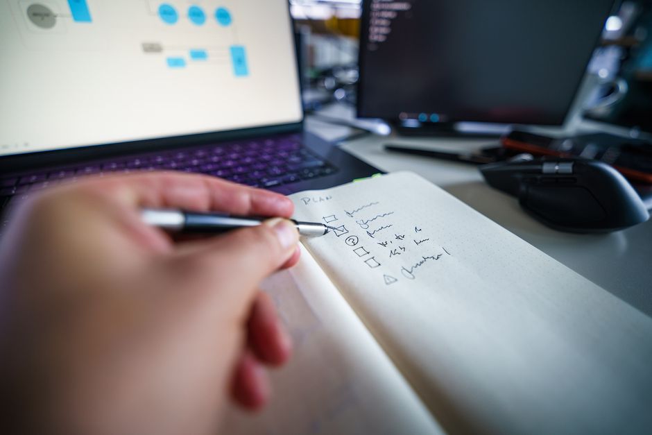 Hand writing in notebook on a desk with laptop, showcasing productivity and organization skills.